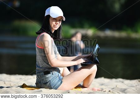 Woman With A Tablet At Beach Sitting In T-shirt And Sexy Denim Shorts On Sand And Looking At Screen.