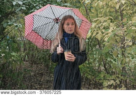 Girl's Waiting For Her Boyfriend With Umbrella And Cup Of Coffee In The Park Under A Heavy Rain In C