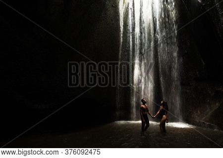 Beautiful Couple In A Cave With A Waterfall. Athletic Man And Woman Under The Streams Of A Waterfall