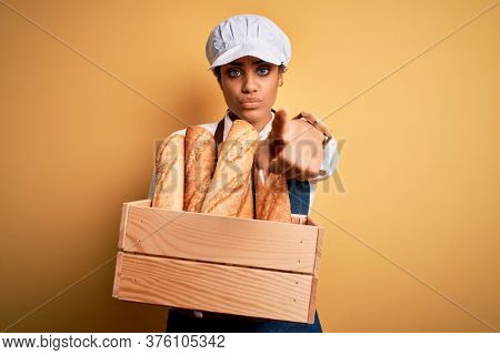 Young african american baker girl wearing apron holding wooden box with bread pointing with finger to the camera and to you, hand sign, positive and confident gesture from the front