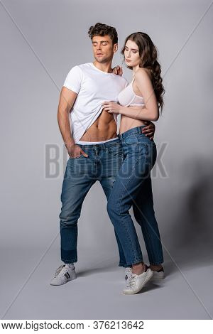 Attractive Woman Touching White T-shirt Of Muscular Boyfriend Standing With Hand In Pocket On Grey
