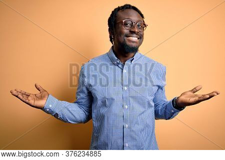 Young handsome african american man wearing shirt and glasses over yellow background smiling showing both hands open palms, presenting and advertising comparison and balance