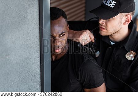 Selective Focus Of Police Officer In Uniform And Cap Holding Truncheon While Arresting African Ameri