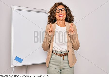 Middle age senior business woman standing on seminar presentation by magnectic blackboard gesturing finger crossed smiling with hope and eyes closed. Luck and superstitious concept.