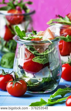 Salad With Arugula, Cheese, Cherry Tomatoes And Prosciutto In Glass Jars, Pink Blue Background, Sele