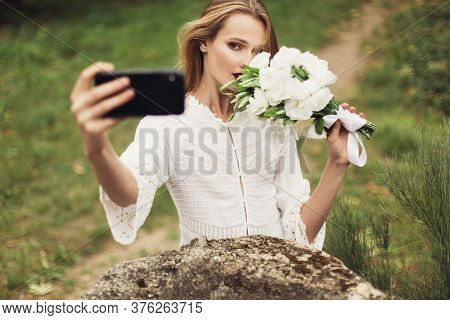 Young Gorgeous Woman In White Dress Holding Little Bouquet Of Flowers Near Face Dreamily Looking In 