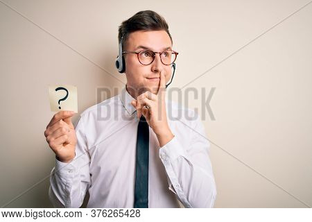 Young call center operator man wearing headset holding paper note with question mark serious face thinking about question, very confused idea