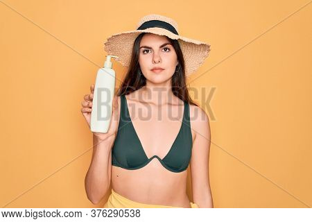 Young beautiful girl wearing swimwear bikini and summer hat holding sun protection cream with a confident expression on smart face thinking serious