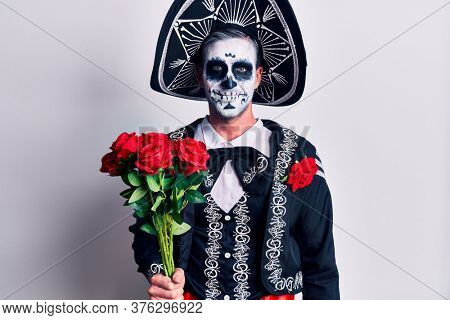 Young man wearing mexican day of the dead costume holding flowers looking positive and happy standing and smiling with a confident smile showing teeth 