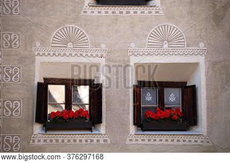 Two Old Wooden Windows With Decorations And Red Geraniums In The Ancient Village Of Guarda, Scuol Mu