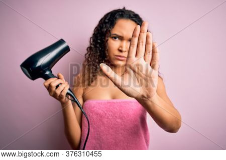 Beautiful woman with curly hair wearing shower towel after bath using hair dryer with open hand doing stop sign with serious and confident expression, defense gesture