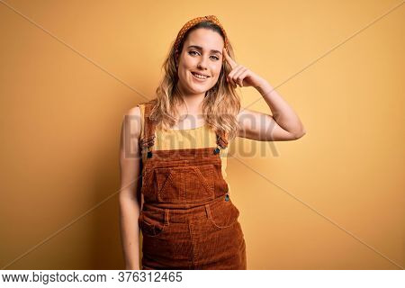 Young beautiful blonde woman wearing overalls and diadem standing over yellow background Smiling pointing to head with one finger, great idea or thought, good memory