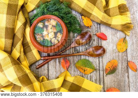 Autumn Transparent Soup With Croutons And Dill In A Clay Pot Against A Yellow Scarf, An Old Wooden B