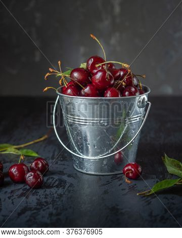 A Bucket Of Cherries On A Dark Background. Berries And Leaves Of Scheresh Are Scattered Around A Tin