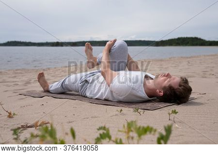 Young Man Practicing Yoga Pavanamuktasana Outdoors On The Beach