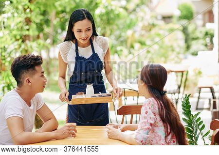 Beautiful Smiling Young Asian Waitress Bringing Tray With Breakfast To Young Happy Couple Sitting At