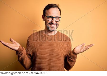 Middle age hoary man wearing brown sweater and glasses over isolated yellow background smiling showing both hands open palms, presenting and advertising comparison and balance