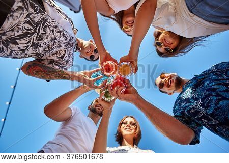 Low Angle View Of Friends Having Fun At Poolside Summer Party, Clinking Glasses With Colorful Summer
