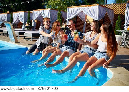Group Of Friends Having Fun At Poolside Party Clinking Glasses With Fresh Cocktails Sitting By Swimm
