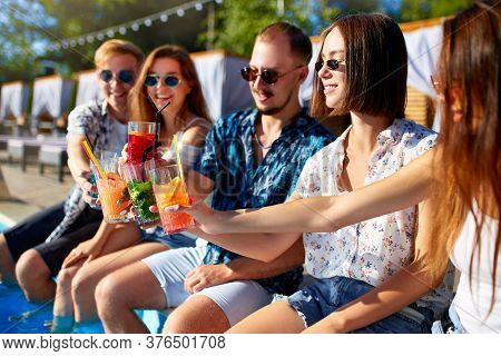 Group Of Friends Having Fun At Poolside Party Clinking Glasses With Fresh Cocktails Sitting By Swimm