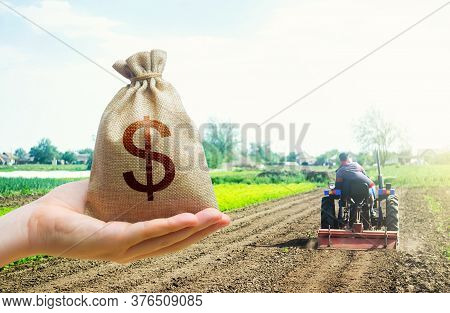 A Hand Holds Out A Dollar Money Bag On A Background Of A Field Cultivating Tractor. Lending Farmers 
