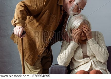 Senior Man With Walking Stick Kissing Head Of Diseased Wife Crying And Covering Eyes With Hands