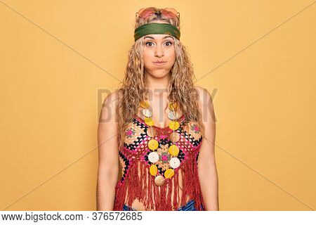 Beautiful blonde hippie woman wearing sunglasses and accessories over yellow background puffing cheeks with funny face. Mouth inflated with air, crazy expression.