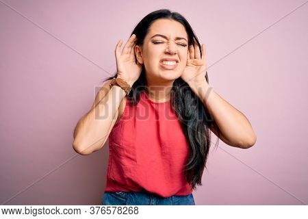 Young brunette woman wearing casual summer shirt over pink isolated background Trying to hear both hands on ear gesture, curious for gossip. Hearing problem, deaf