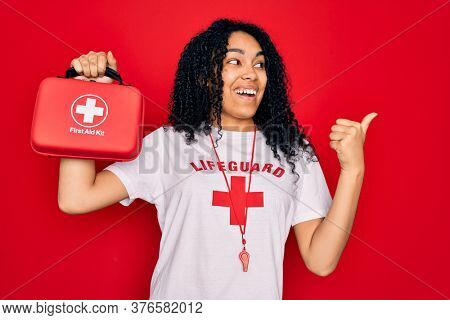Young african american curly lifeguard woman wearing whistle holding first aid kit pointing and showing with thumb up to the side with happy face smiling