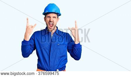 Young handsome man wearing worker uniform and hardhat shouting with crazy expression doing rock symbol with hands up. music star. heavy music concept. 