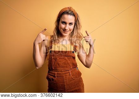 Young beautiful blonde woman wearing overalls and diadem standing over yellow background smiling pointing to head with both hands finger, great idea or thought, good memory