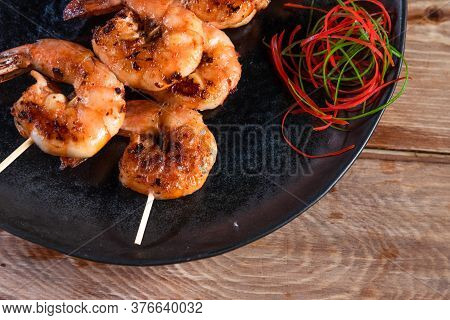 Appetizing Fried Shrimp On A Skewer In A Black Plate On A Wooden Surface Among Lemon And Seasonings.