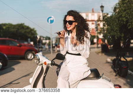 Young Woman With Dark Curly Hair In White Costume And Sunglasses Sitting On White Moped And Emotiona