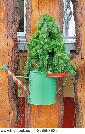 An Artificial Coniferous Tree In A Red-green Watering Can Hanging On A Wooden Fence With A Garland