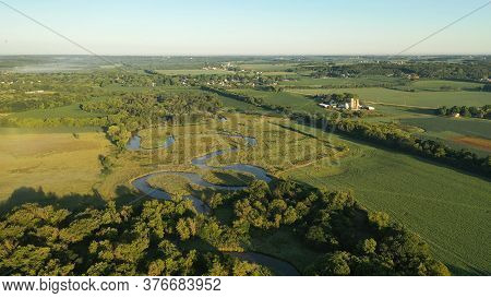 Aerial View Of Rural Landscape With Lush Greenery, Trees, River In The Foreground And Farmhouse, Far