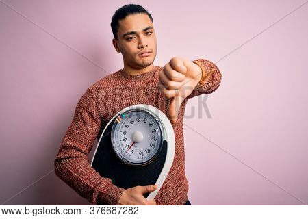 Young brazilian man doing diet to lose weigth holding scale over isolated pink background with angry face, negative sign showing dislike with thumbs down, rejection concept