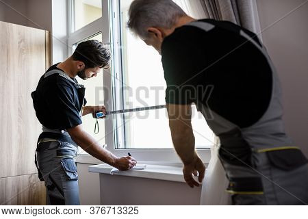 Two Professional Workers In Uniform Using Tape Measure While Measuring Window And Making Notes For I
