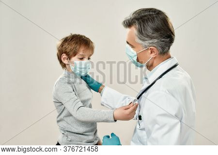 Professional Mature Doctor And Little Patient In Protective Masks. Male Pediatrician Working With Li