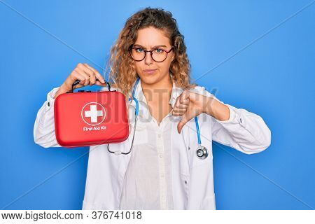 Young beautiful blonde doctor woman with blue eyes wearing stethoscope holding first aid kit with angry face, negative sign showing dislike with thumbs down, rejection concept