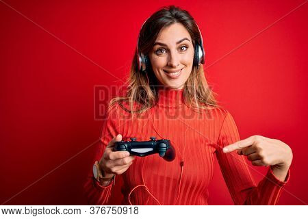 Young beautiful brunette gamer woman playing video game using joystic and headphones very happy pointing with hand and finger