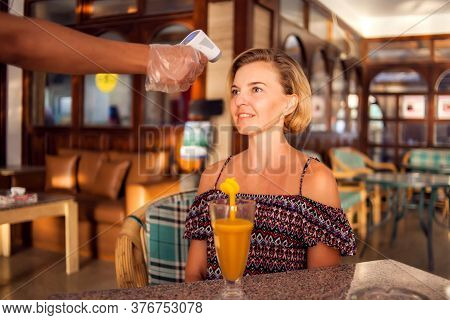 A Worker Measuring Temperature Of Woman Visitior Inside  Of The Cafe. Life During Coronavirus Time