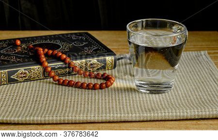 Glass Of Clear Water, Stone Rosary And Green And Gilded Book Of Quran On A Napkin. First Drink, Musl