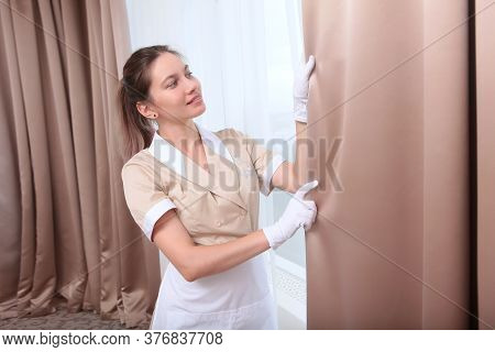 A Maid In White Gloves And Uniform Adjusts The Curtains In A Hotel Room Or In A House. The Concept O