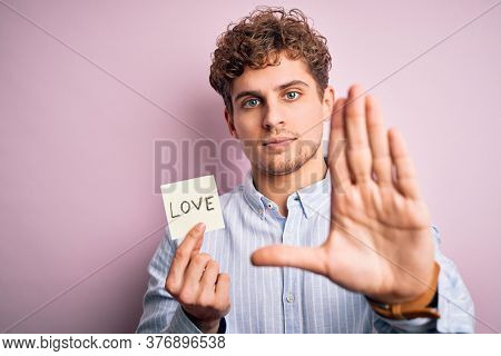 Young blond romantic man with curly hair holdiing paper with love message for valentines day with open hand doing stop sign with serious and confident expression, defense gesture