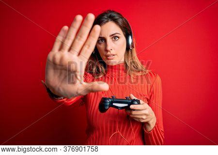 Young beautiful brunette gamer woman playing video game using joystic and headphones with open hand doing stop sign with serious and confident expression, defense gesture