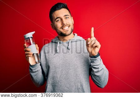 Young handsome sportsman drinking bottle of water to refeshment over red background surprised with an idea or question pointing finger with happy face, number one