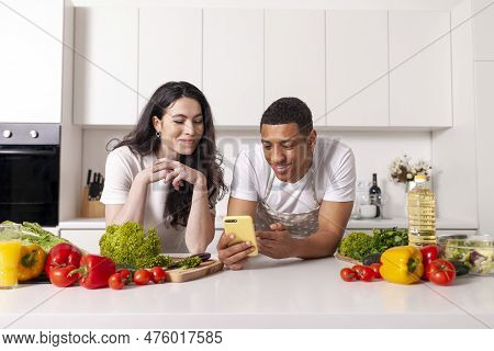 Young Multiracial Couple Looking At Phone And Checking Grocery List In White Modern Kitchen