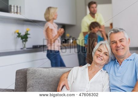 Älteres Ehepaar auf der Couch sitzen und Lächeln in die Kamera, während ihre Familie in die Kitc steht