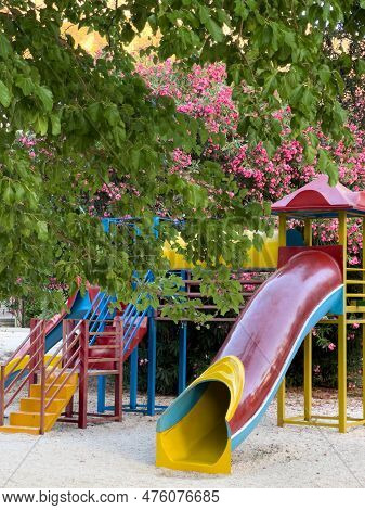 Slide-pipe With A Cover And Steps Stands On A Playground Near Flowering Trees