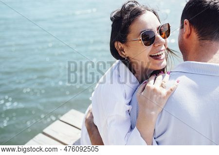 Young Couple In Love In Light Casual Clothes Is Sitting On Wooden Bridge. Happy Woman And Man Hug An